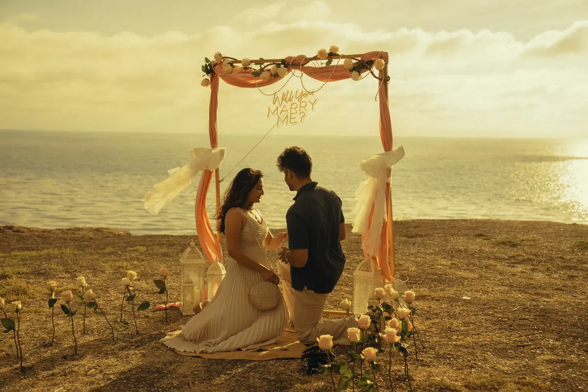 Nihal on one knee under a flower arch, proposing to Rakshana by the sea.